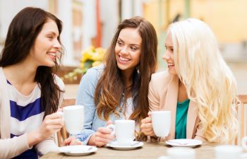 Three women are sitting at an outdoor table, holding cups of coffee and smiling at each other. They are engaged in conversation.