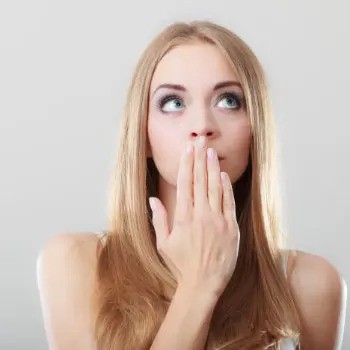 A blonde woman with long straight hair covers her mouth with her hand and looks upward with a thoughtful expression. She is set against a plain gray background.