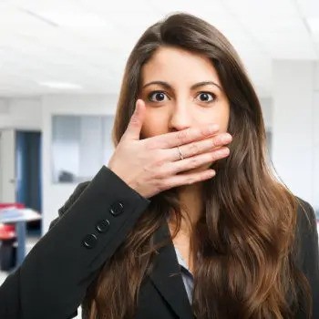 A woman with long brown hair covers her mouth with one hand while standing in an office setting.