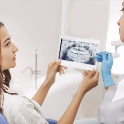 A person in a dentist's chair is holding an X-ray image while a dental professional points to it, discussing the scan.