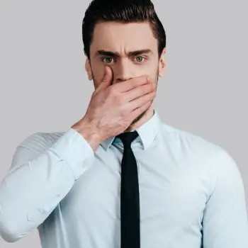 A man in a light blue shirt and black tie covers his mouth with one hand, looking directly at the camera with a concerned expression.