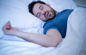 A person sleeping in bed, wearing a blue shirt, with their mouth open and arm resting on the pillow.