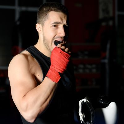 Boxing fighter with red hand wraps is putting in a mouthguard in a dimly lit gym.