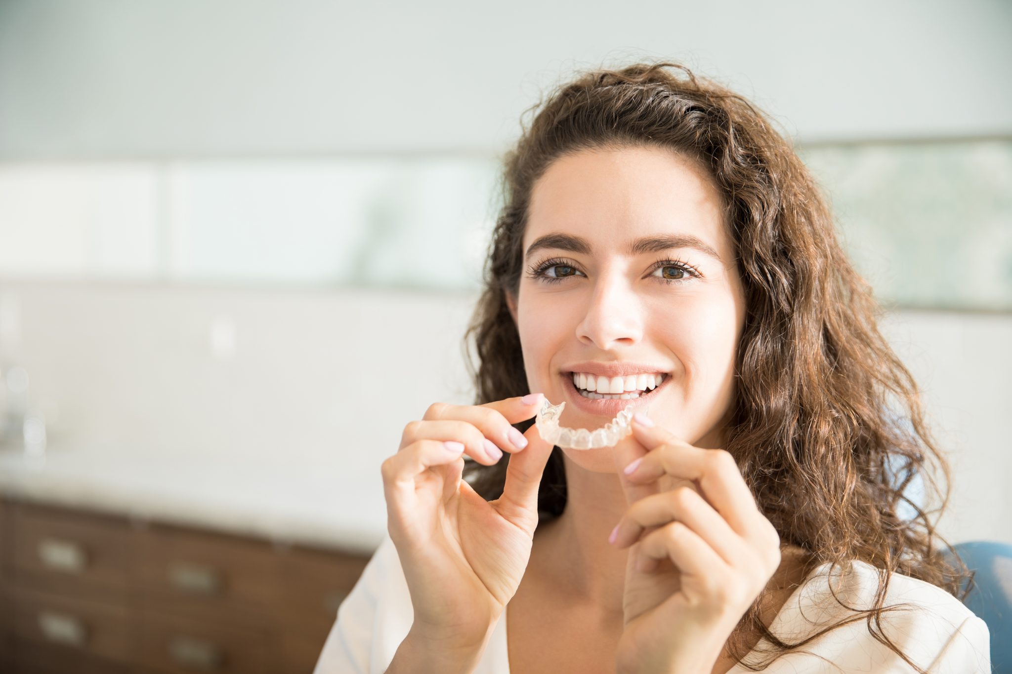 Person smiling while holding a clear dental retainer in an indoor setting.