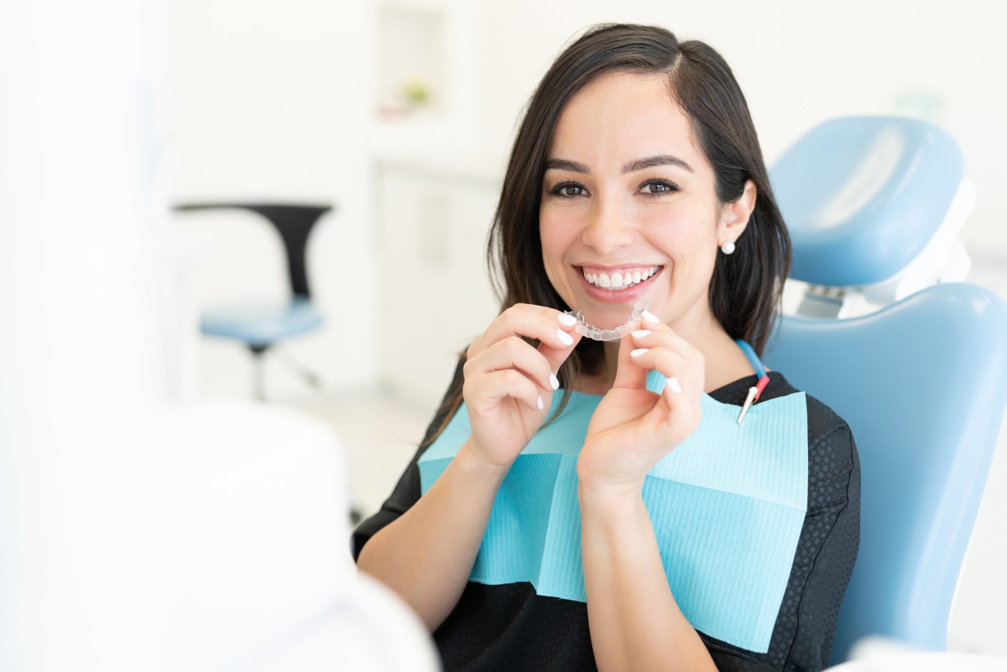 A woman with dark hair is sitting in a dentist's chair, smiling and holding a clear dental aligner. She is wearing a blue dental bib.