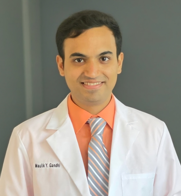 Dr. Maulik Gandhi, DDS, MPH, MBA — a man wearing a white lab coat, orange shirt, and striped tie stands in front of a gray background, smiling at the camera.